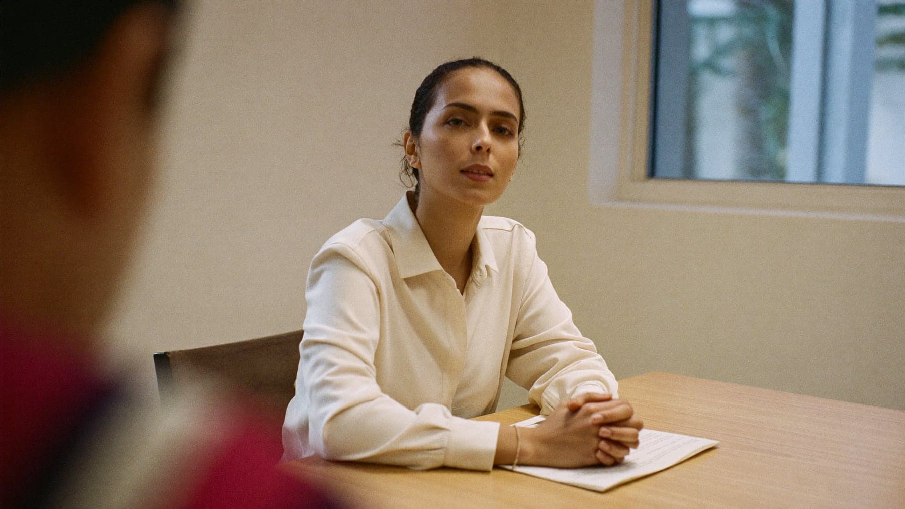 A young professional mid-answer in an interview, composed, her printed résumé on the table in front of her.